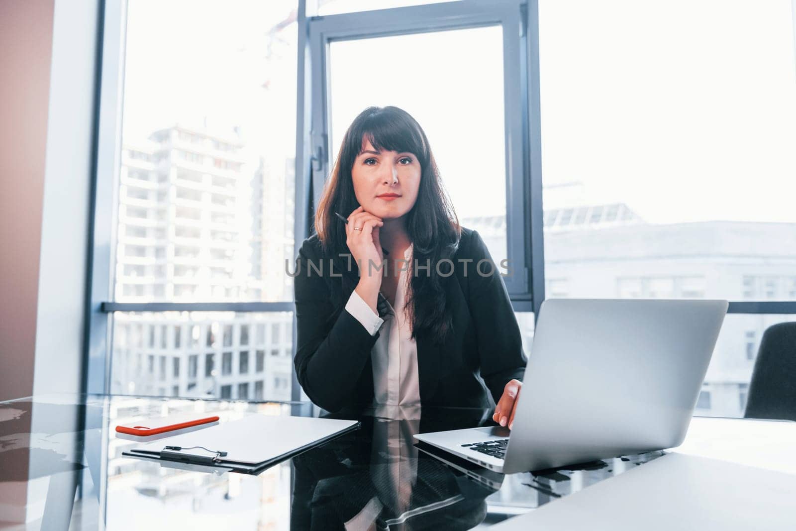 With laptop on table. Woman in black formal clothes is indoors in the modern office.