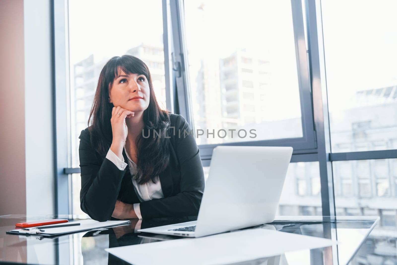 With laptop on table. Woman in black formal clothes is indoors in the modern office.