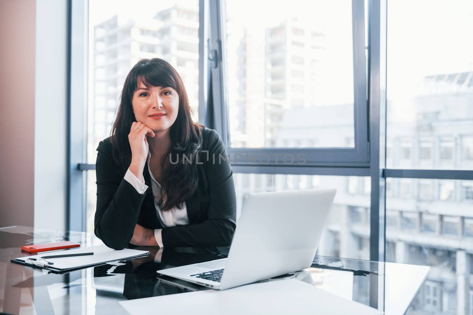 With laptop on table. Woman in black formal clothes is indoors in the modern office.