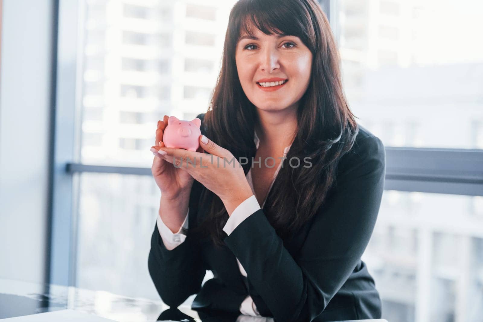 Woman in black formal clothes is indoors in the modern office holds pink piggy bank.