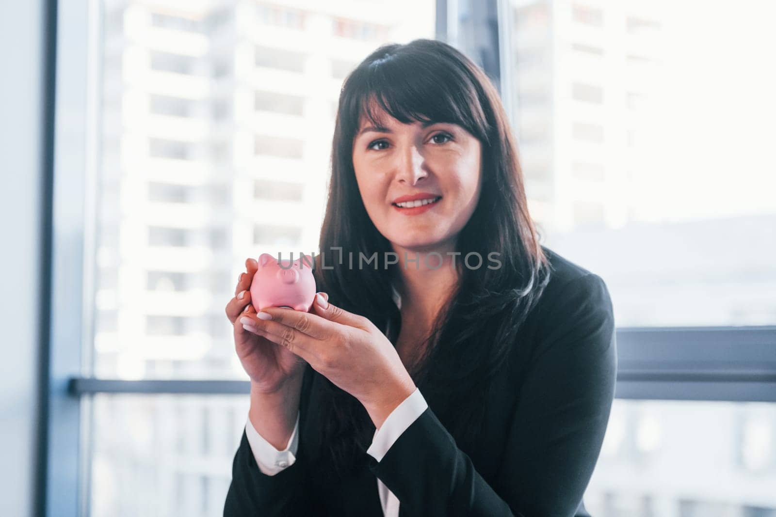 Woman in black formal clothes is indoors in the modern office holds pink piggy bank.