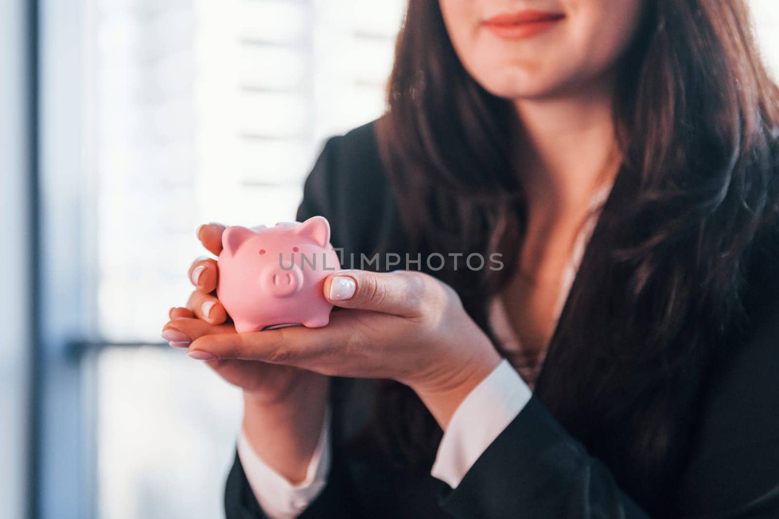 Woman in black formal clothes is indoors in the modern office holds pink piggy bank.