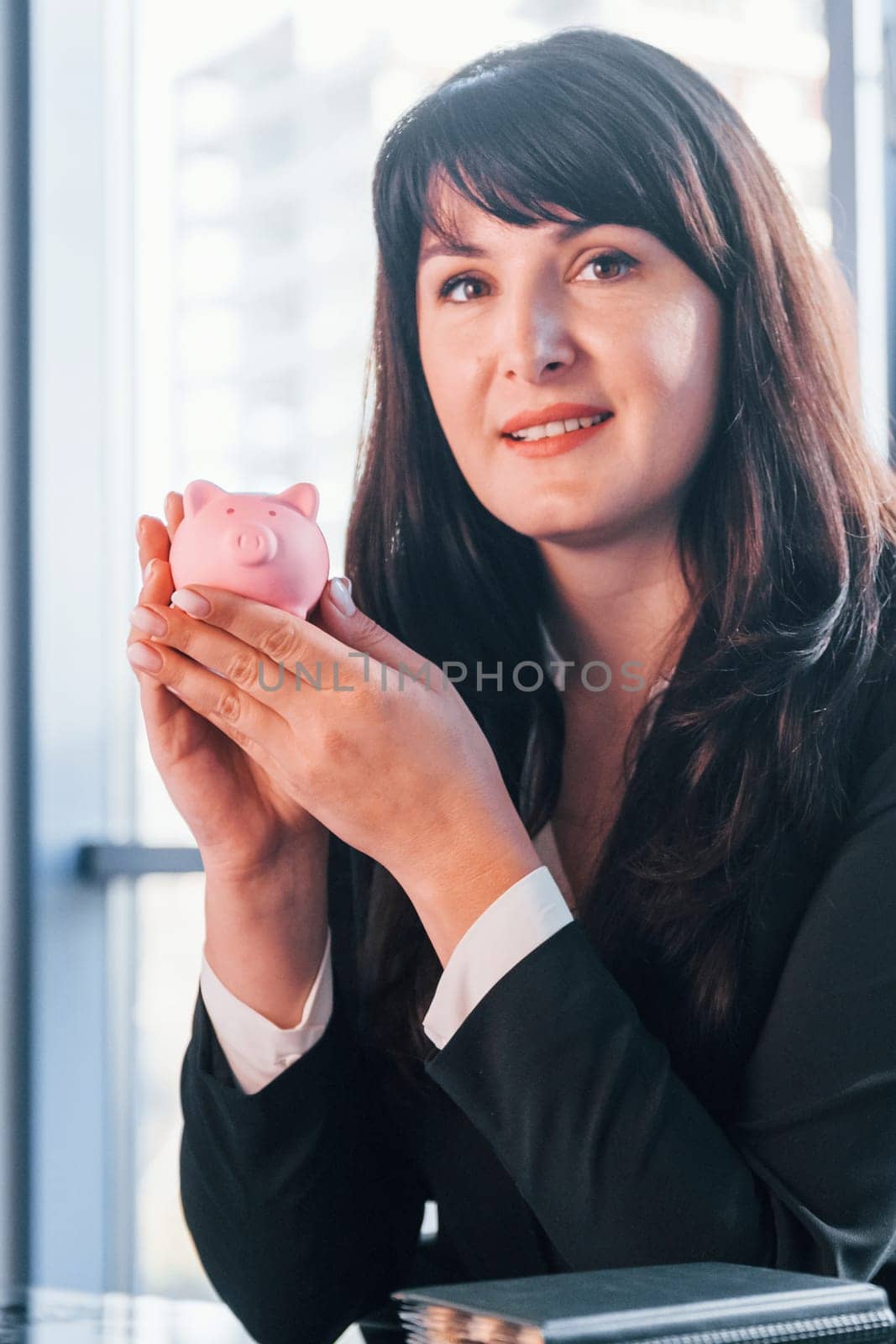 Woman in black formal clothes is indoors in the modern office holds pink piggy bank.