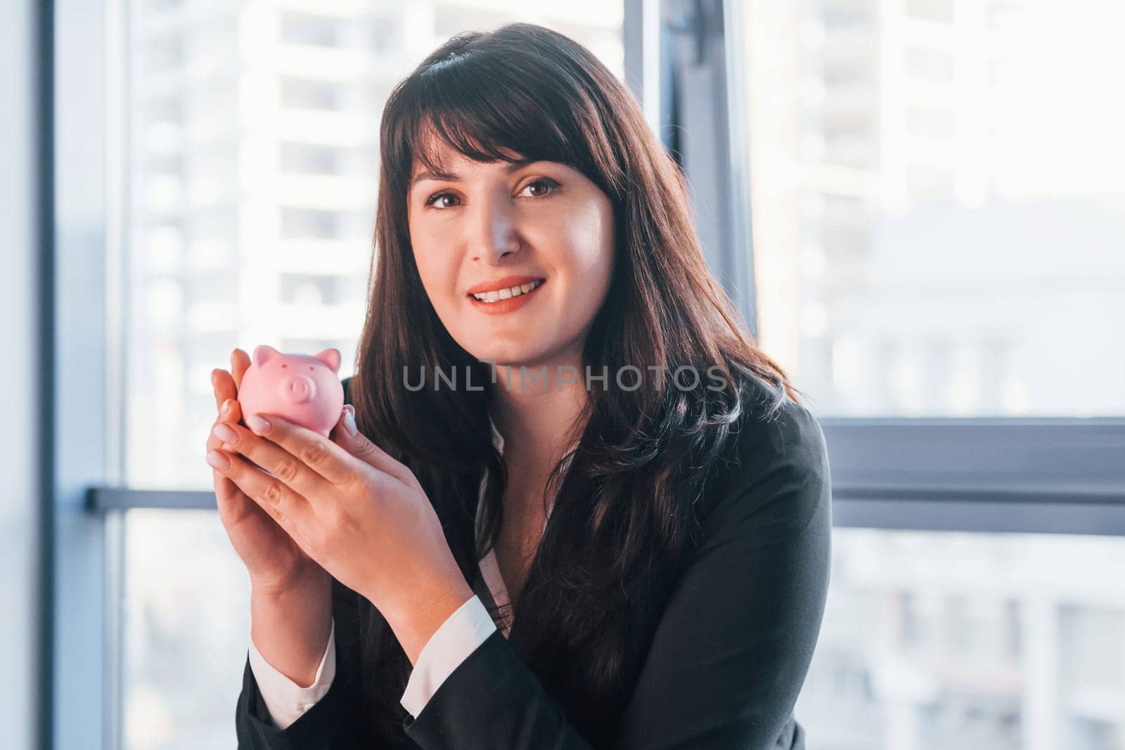 Woman in black formal clothes is indoors in the modern office holds pink piggy bank.