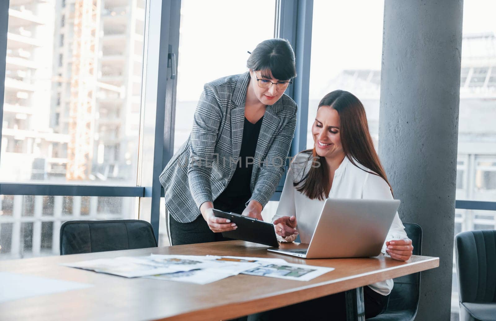 Using laptop. Two women in formal clothes is indoors in the modern office works together.