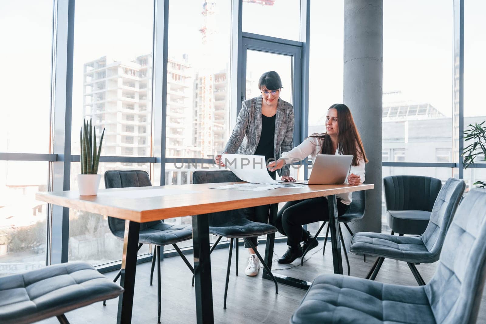 Using laptop. Two women in formal clothes is indoors in the modern office works together.