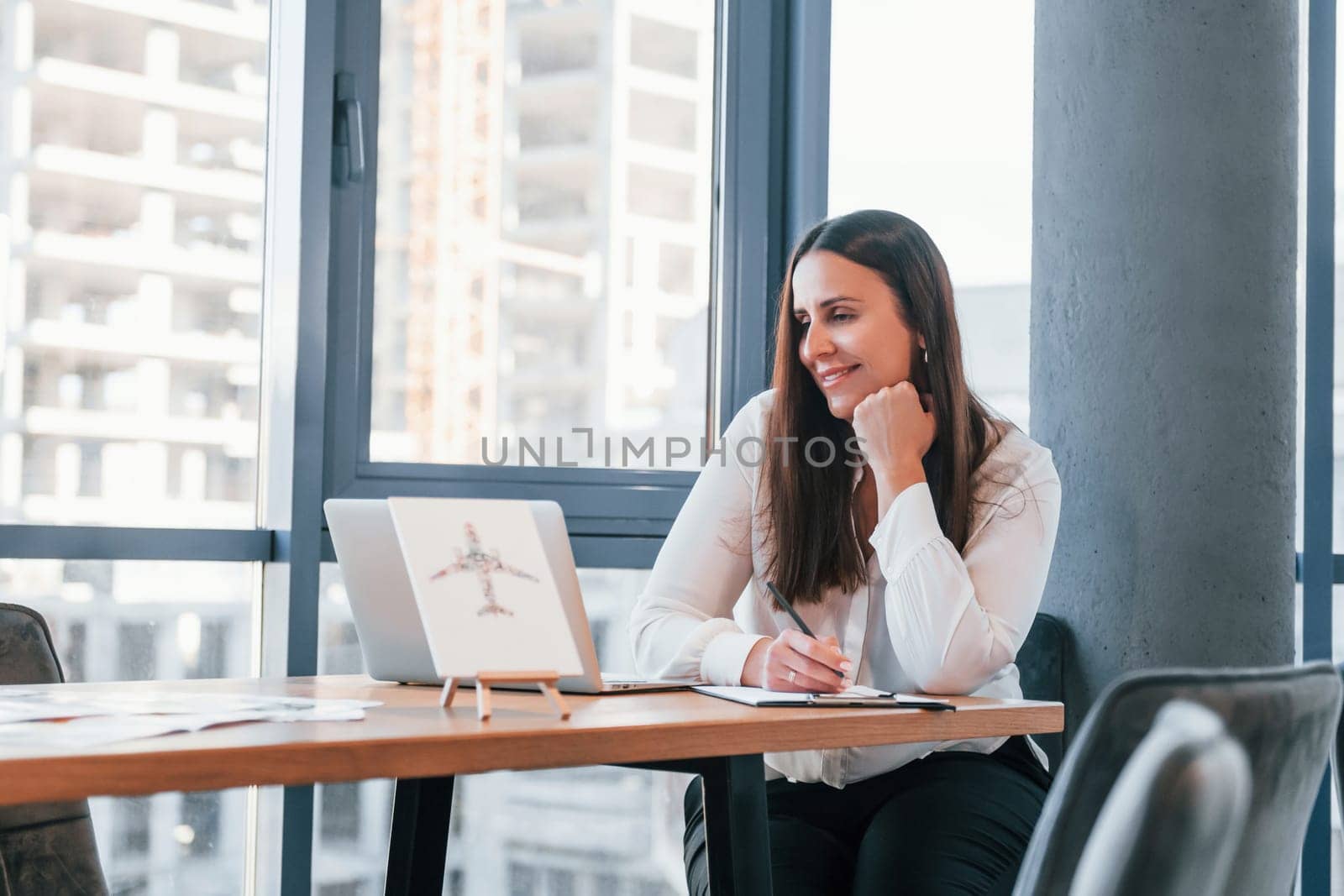 Sits by the table. Young woman in white formal clothes is indoors in the modern office.