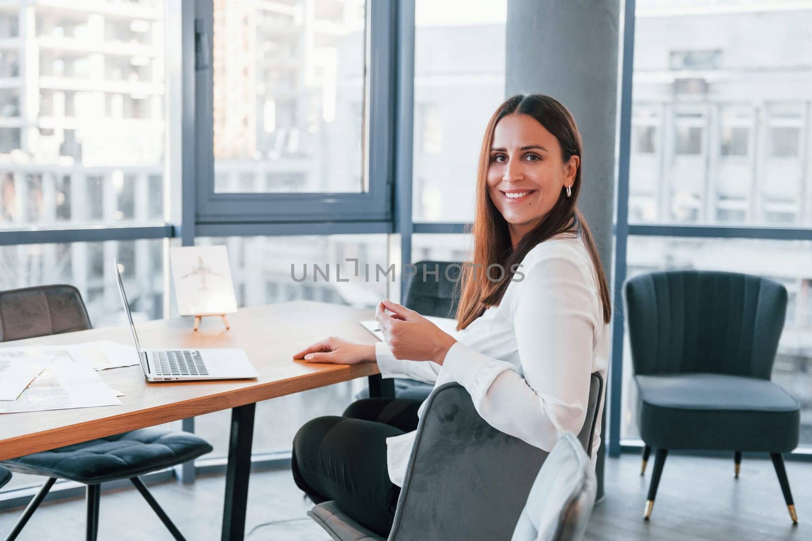 Sits by the table. Young woman in white formal clothes is indoors in the modern office.