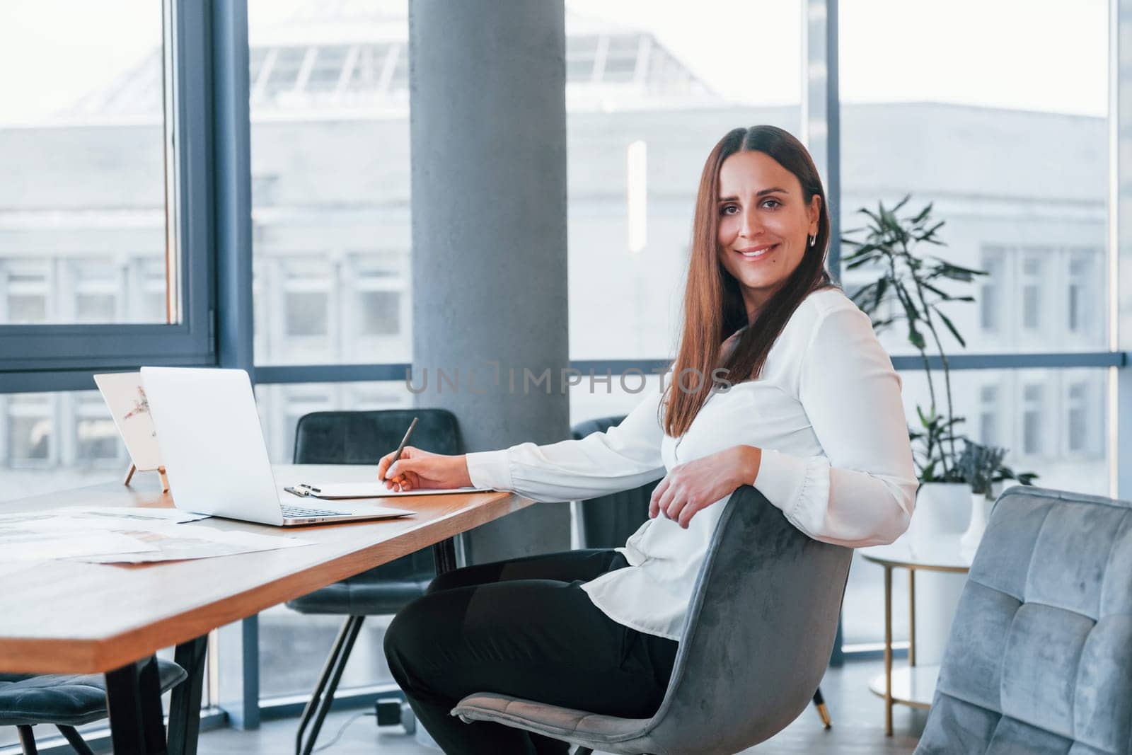 Sits by the table. Young woman in white formal clothes is indoors in the modern office.