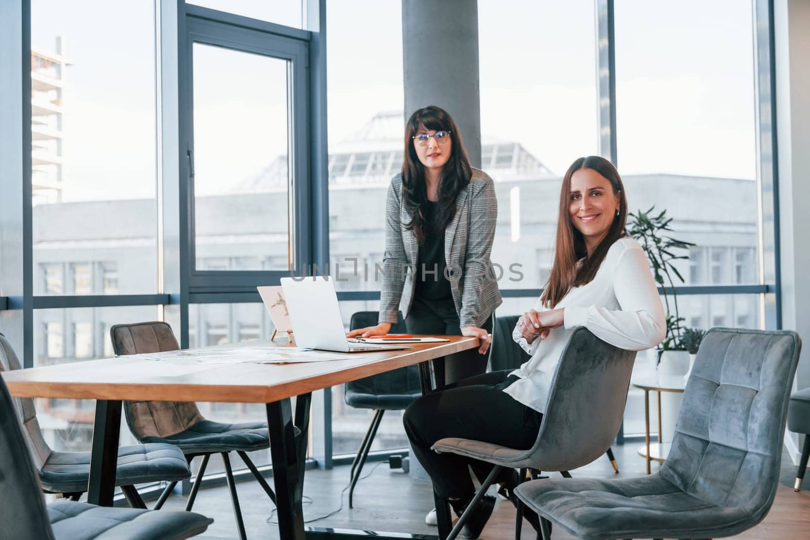 Two women in formal clothes is indoors in the modern office works together.