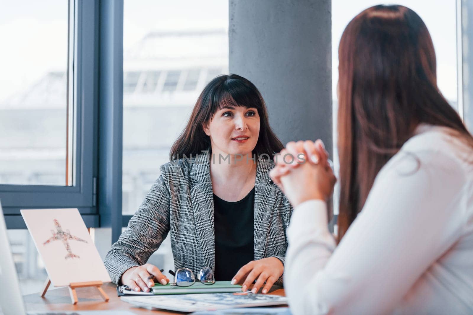 Sitting by the table. Two women in formal clothes is indoors in the modern office works together.