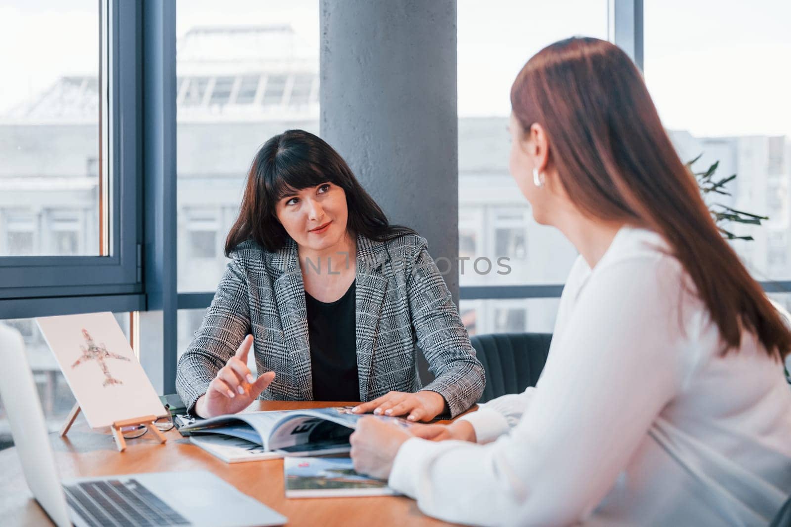 Sitting by the table. Two women in formal clothes is indoors in the modern office works together.