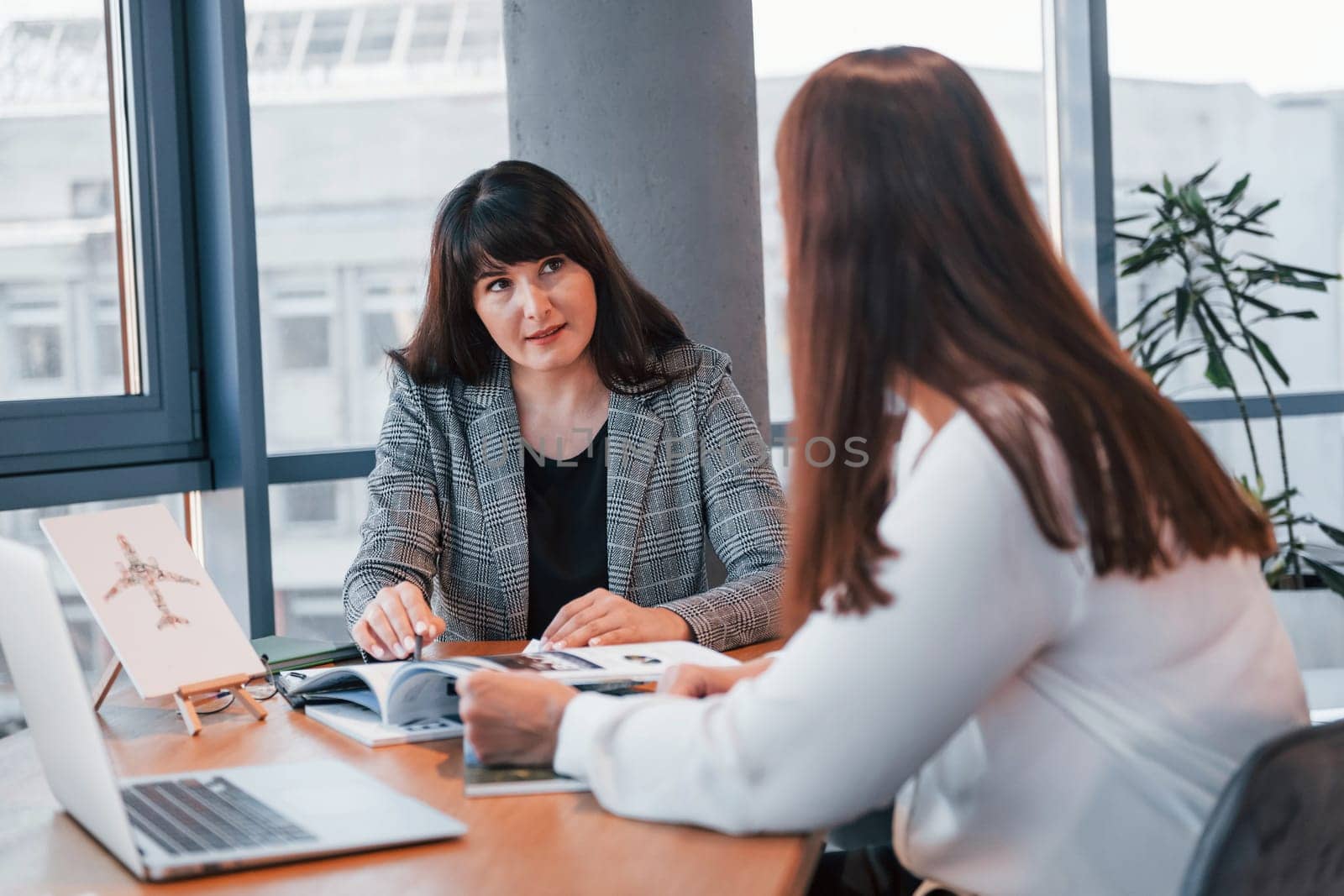 Sitting by the table. Two women in formal clothes is indoors in the modern office works together.