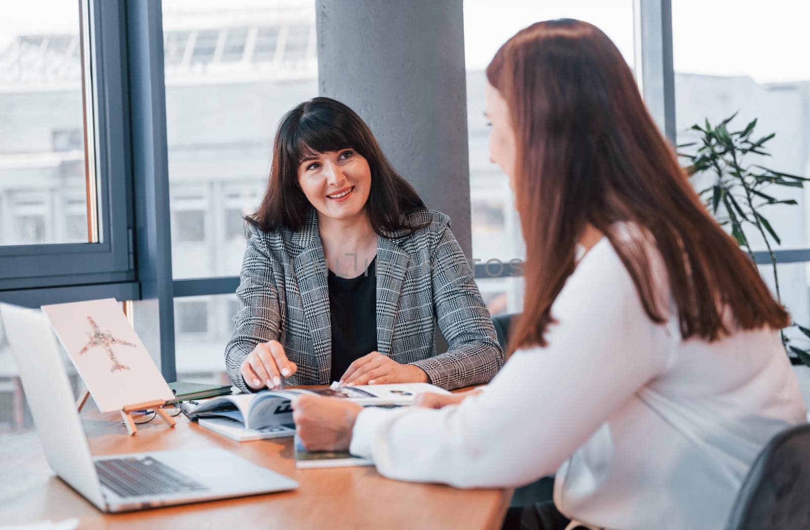 Sitting by the table. Two women in formal clothes is indoors in the modern office works together.