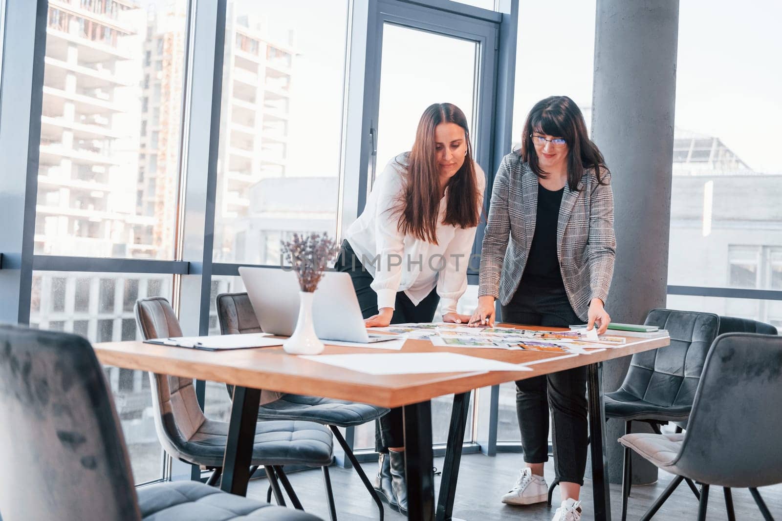 Standing and talking. Two women in formal clothes is indoors in the modern office works together.