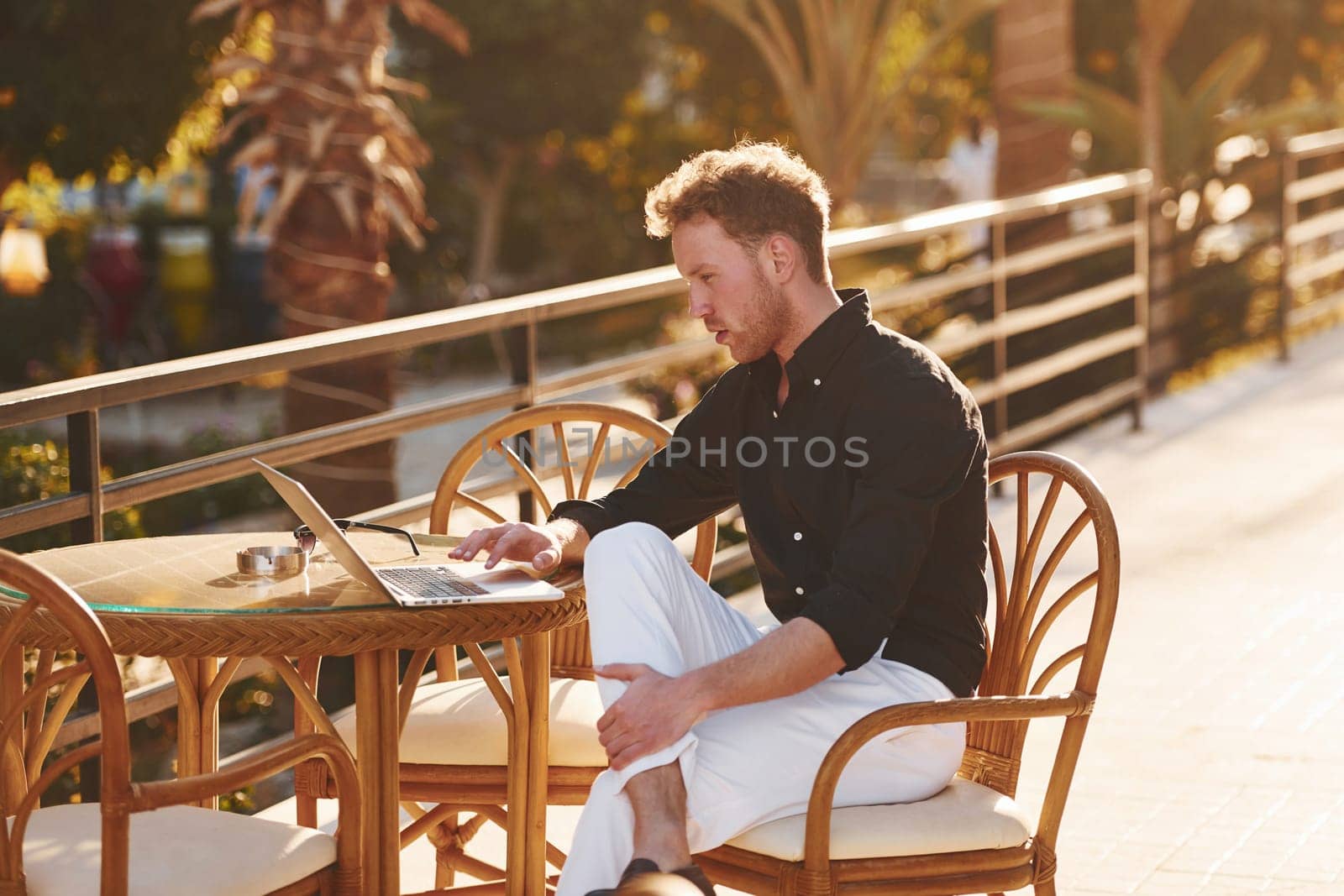 Sits by the table. Young man is outdoors at sunny daytime. Concept of vacation.