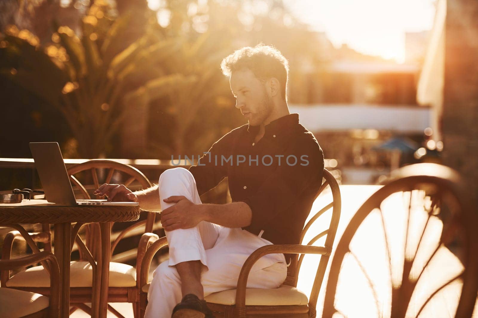 Sits by the table. Young man is outdoors at sunny daytime. Concept of vacation.