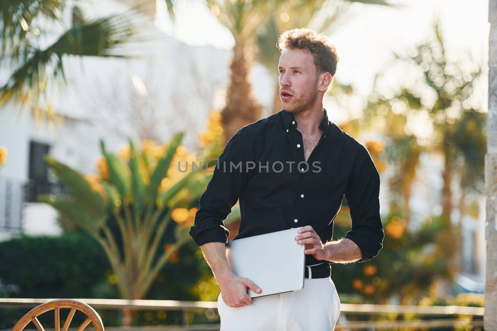 Holds laptop. Young man is outdoors at sunny daytime. Concept of vacation.