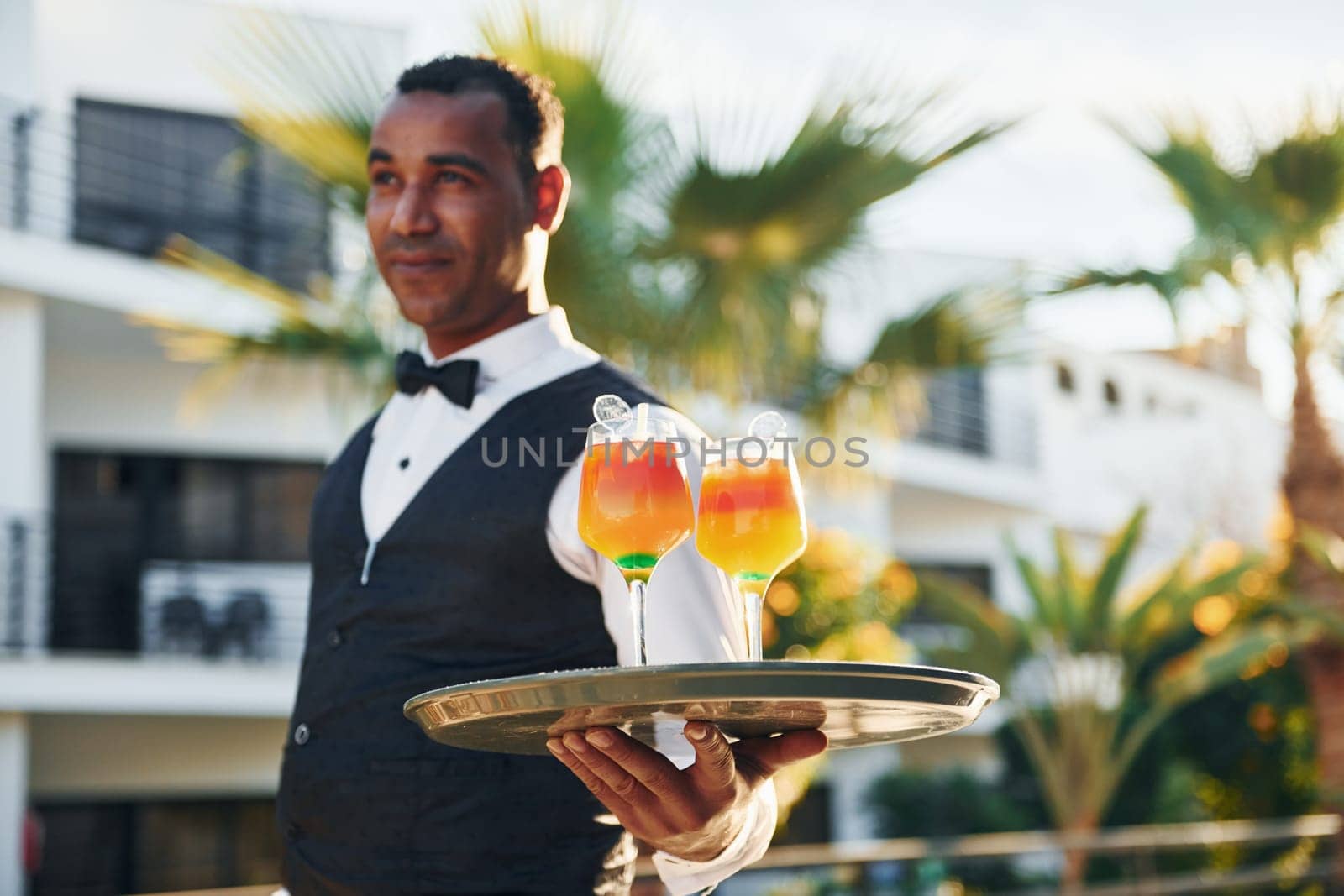 Holds cocktails. Black waiter in formal clothes is at his work outdoors at sunny daytime.