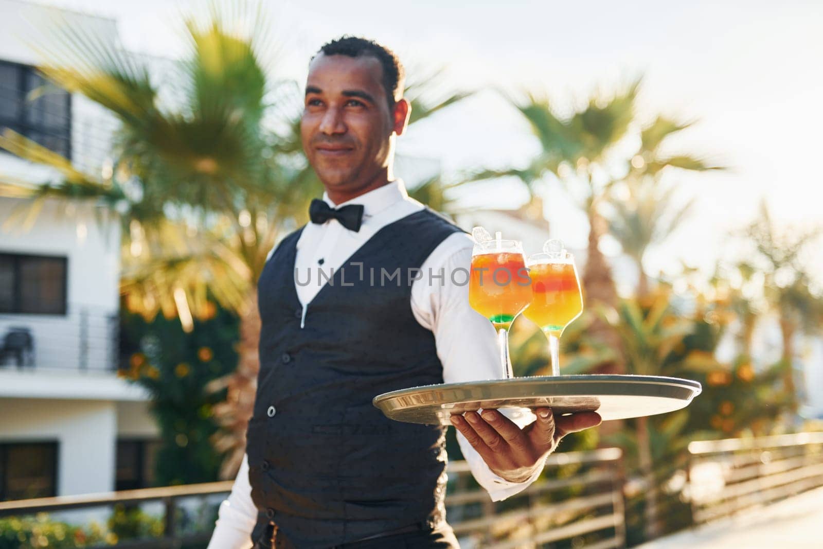 Holds cocktails. Black waiter in formal clothes is at his work outdoors at sunny daytime.