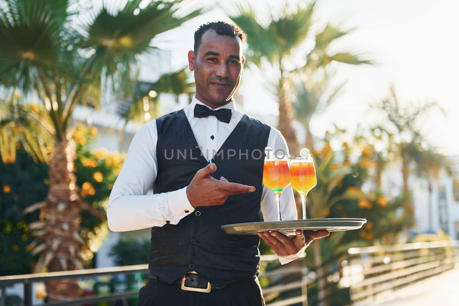 Holds cocktails. Black waiter in formal clothes is at his work outdoors at sunny daytime.