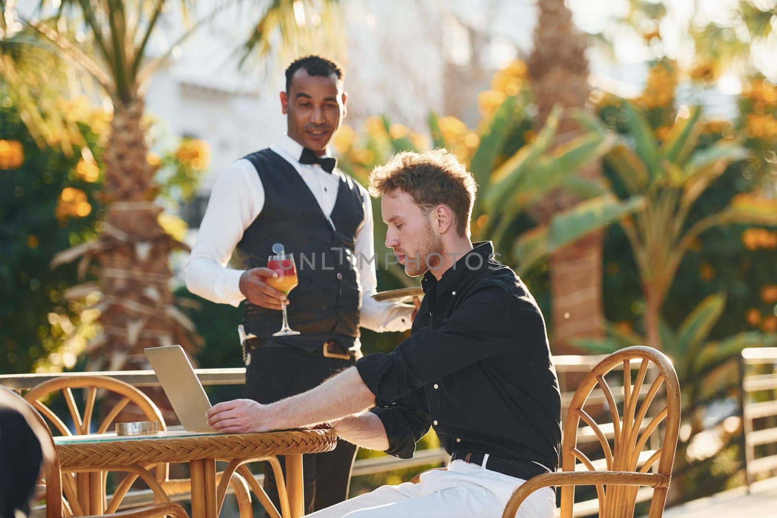 Serviced by waiter. Young man is outdoors at sunny daytime. Concept of vacation.