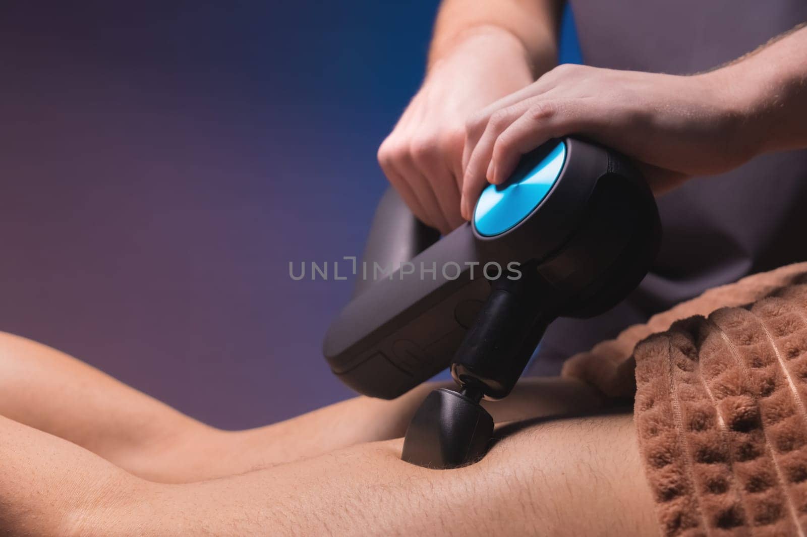 Close-up of the hand of an unrecognizable male massage therapist massaging with a massage gun percussion tool a muscular athlete, in a spa treatment, lying on his back in a massage table.