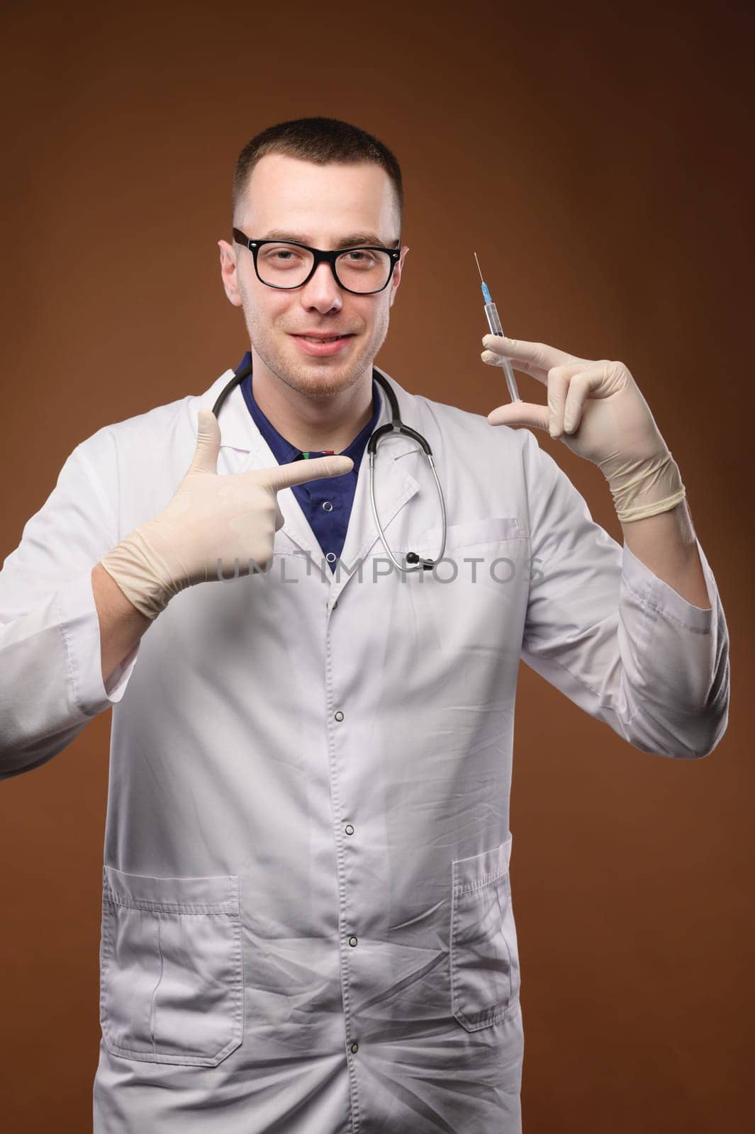 Covid-19, coronavirus disease, medical workers concept. Pleasant smiling male doctor, a doctor in a medical mask and gloves, points with a finger and holds a syringe with a vaccine.