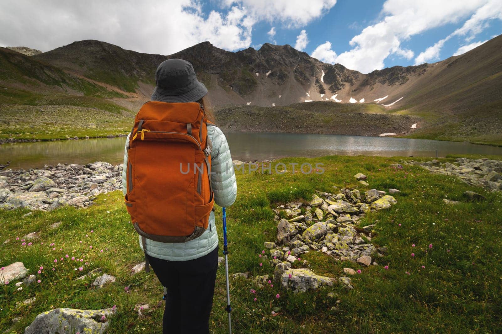 A traveling free man with an orange backpack walks along a path towards a high mountain range and a lake. Dramatic landscape with a tourist high in the mountains.