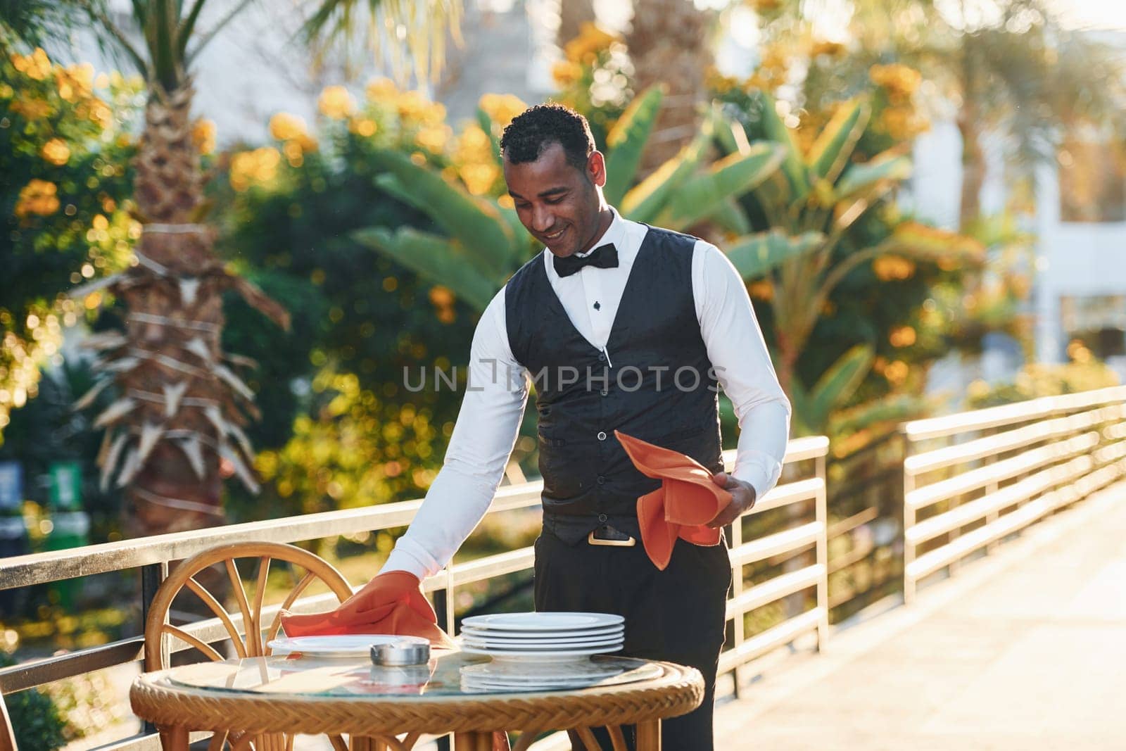 Wipes plates by red cloth. Black waiter in formal clothes is at his work outdoors at sunny daytime.