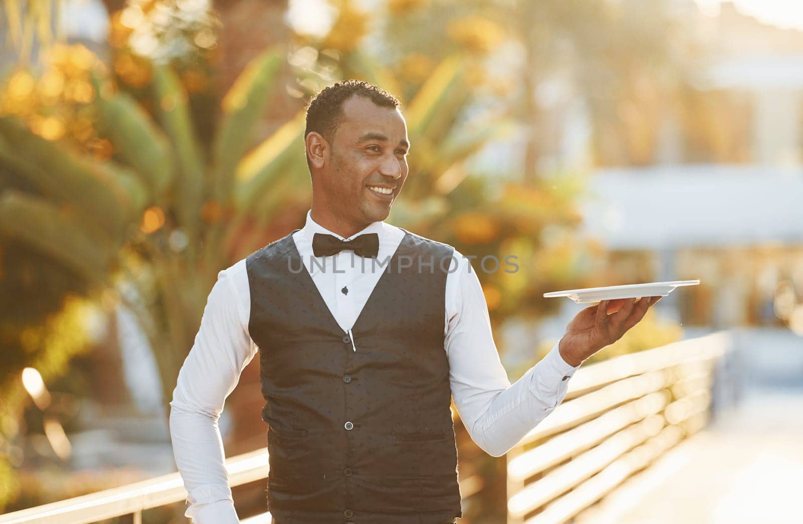 Holds empty plate. Black waiter in formal clothes is at his work outdoors at sunny daytime.