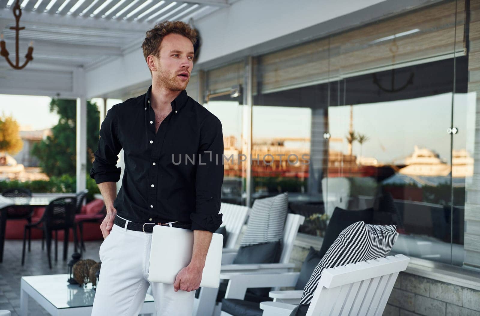 Holds laptop. Young man is outdoors at sunny daytime. Concept of vacation.