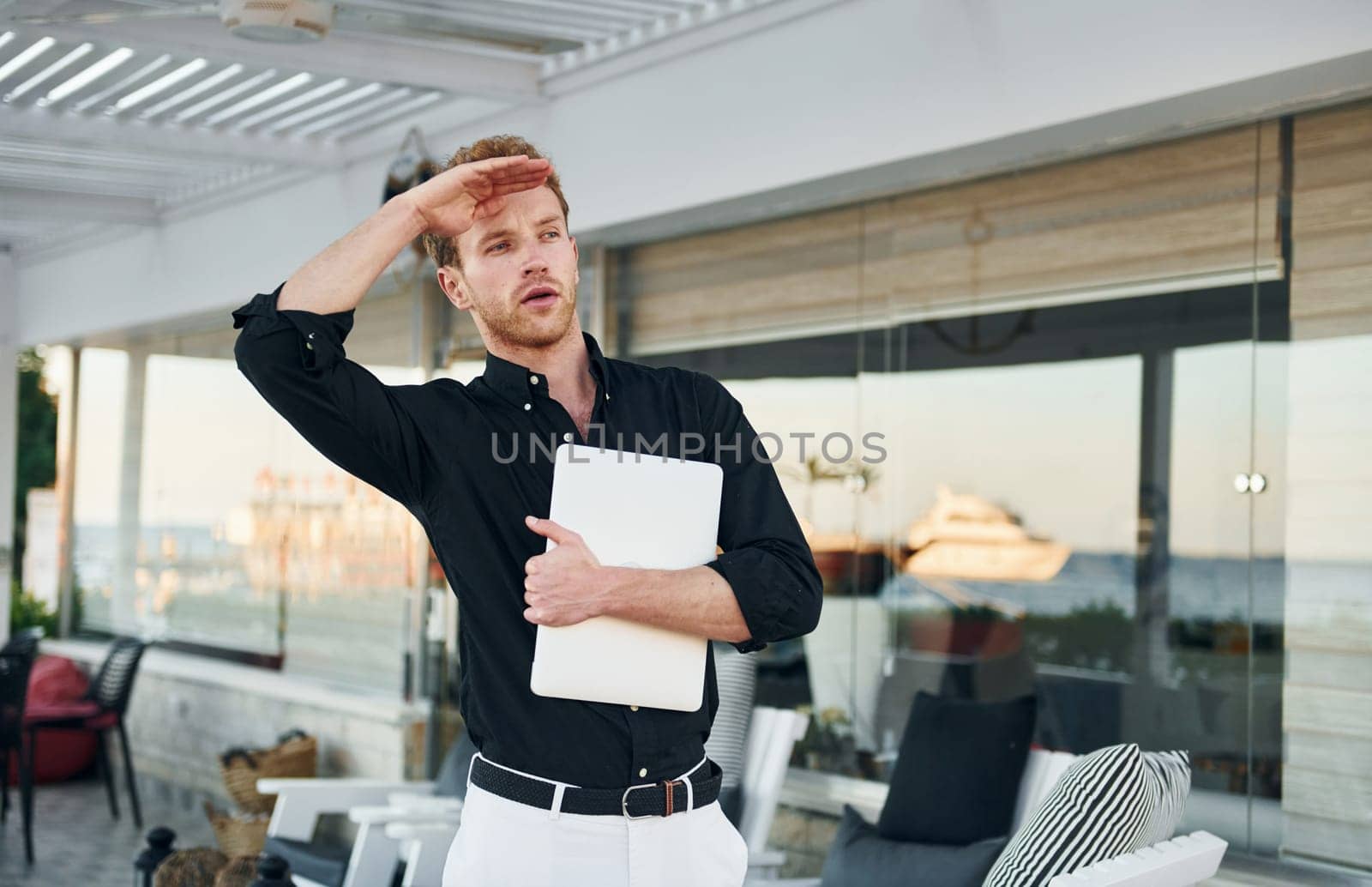 Holds laptop. Young man is outdoors at sunny daytime. Concept of vacation.