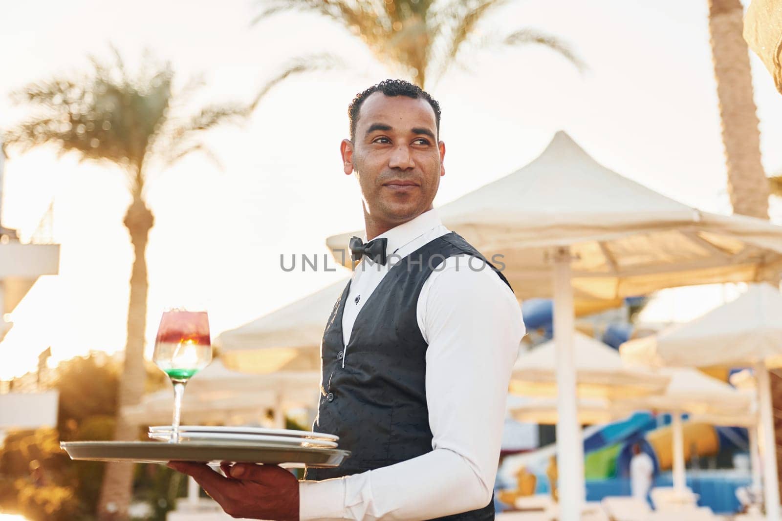 With order in hand. Black waiter in formal clothes is at his work outdoors at sunny daytime.