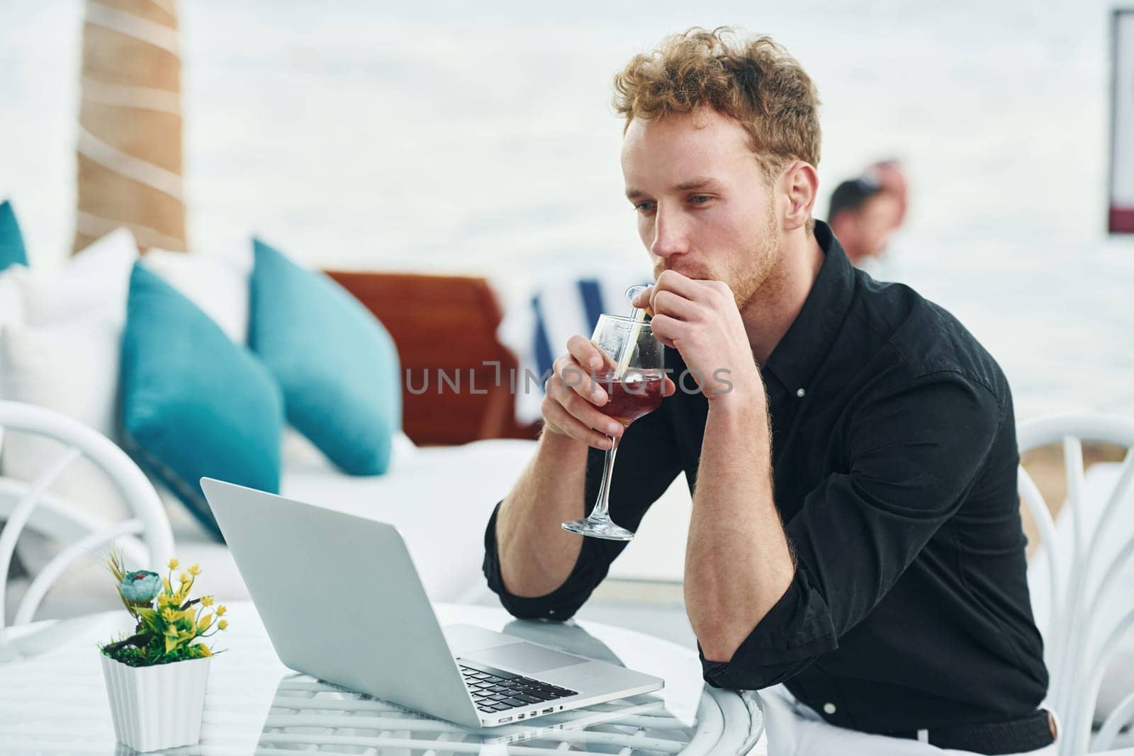 Sits by the table. Young man is outdoors at sunny daytime. Concept of vacation.
