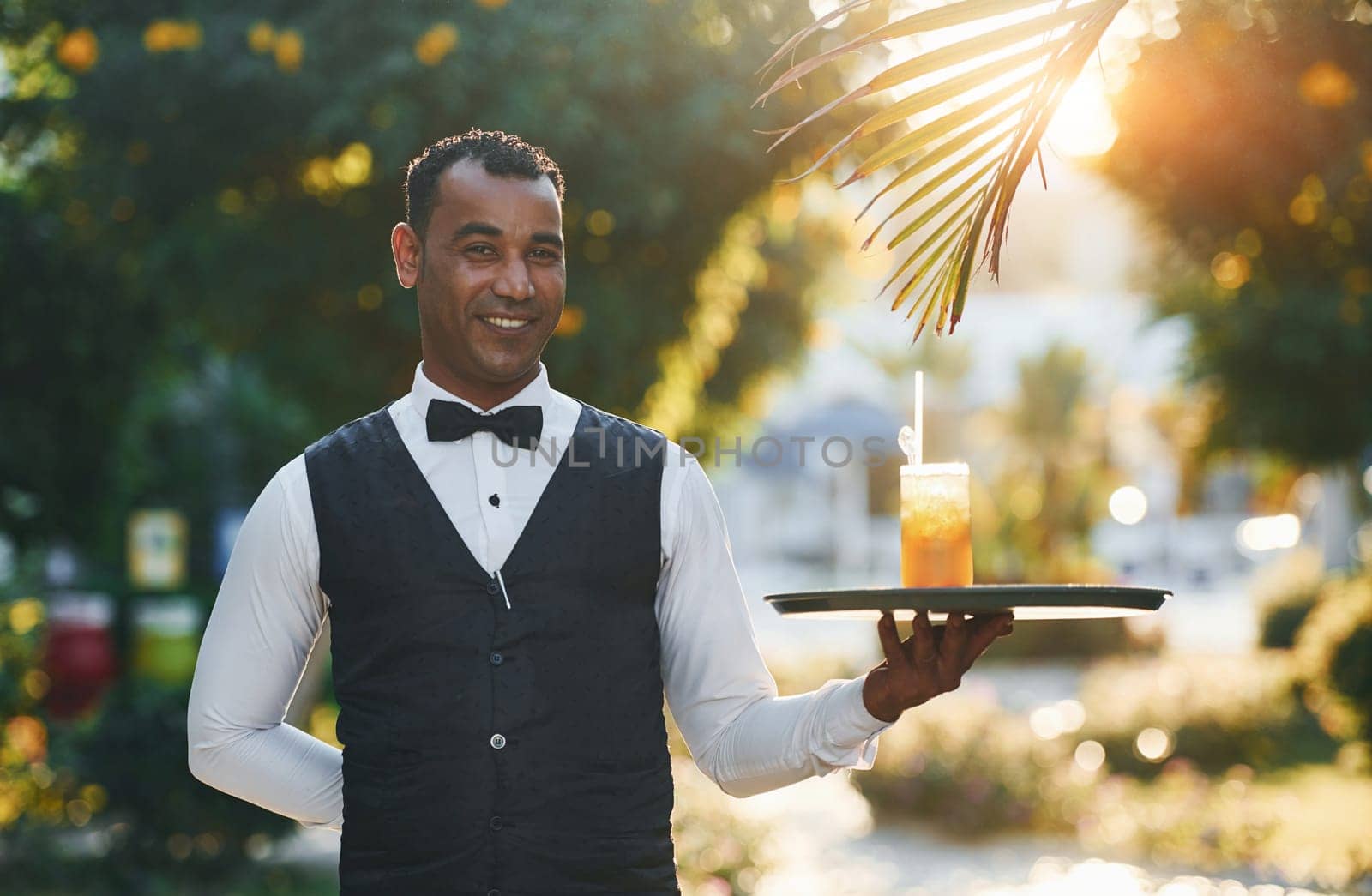 With order in hand. Black waiter in formal clothes is at his work outdoors at sunny daytime.