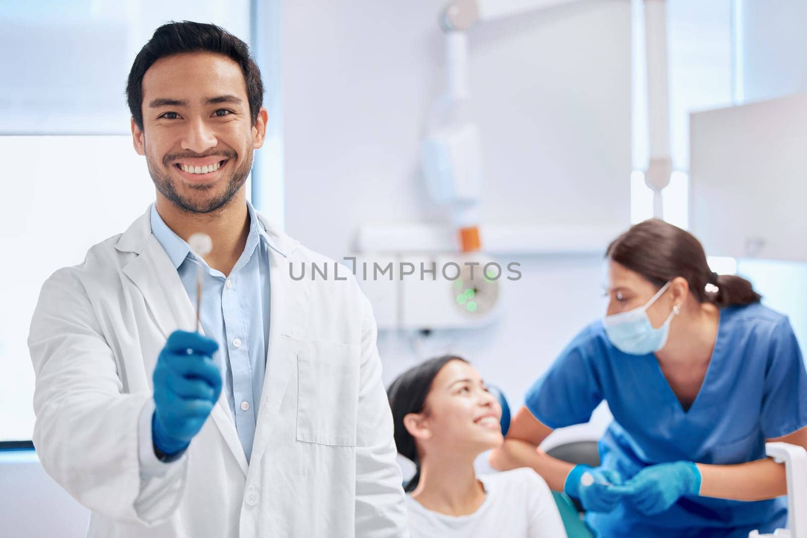 Providing his patients with high grade tools. a young dentist holding up a tool while his assistant helps a patient. by YuriArcurs