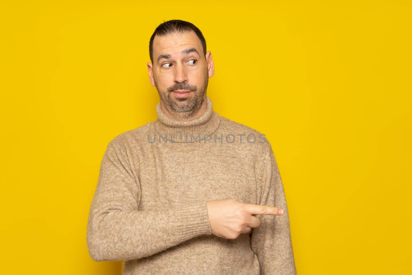 Hispanic man with a beard in a beige turtleneck pointing to the side with his index finger while looking sideways skeptically, he is very curious about what is happening next to him. Isolated