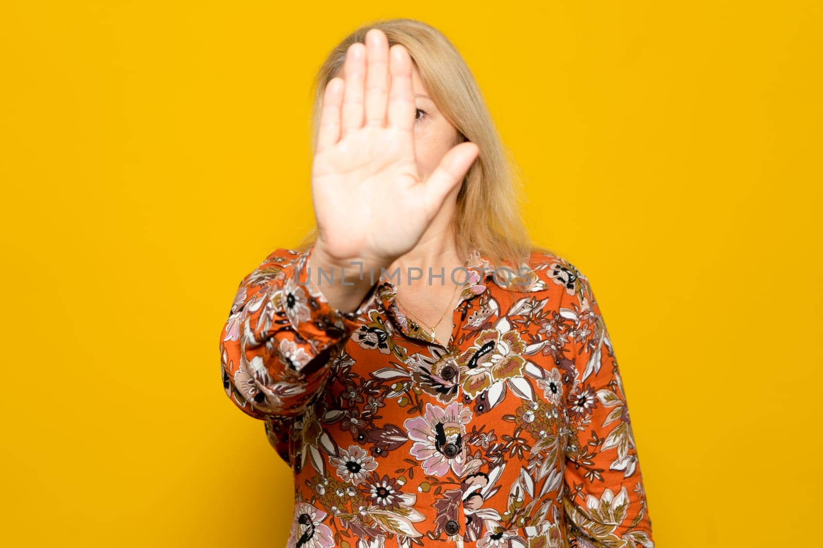 Caucasian blonde woman in a patterned jumper raised her hand in deterrence, campaign to stop violence against women. Woman raised her hand indignantly. Copy space, isolated on yellow background