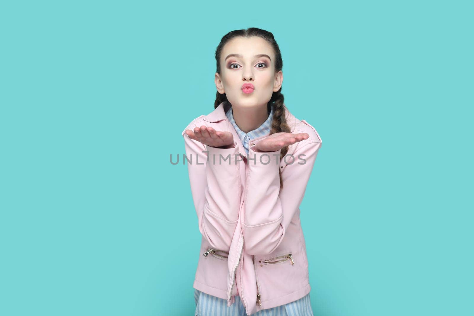 Portrait of romantic flirting teenager girl with braids wearing pink jacket standing expressing love and gentle, sending air kisses over palms. Indoor studio shot isolated on green background.