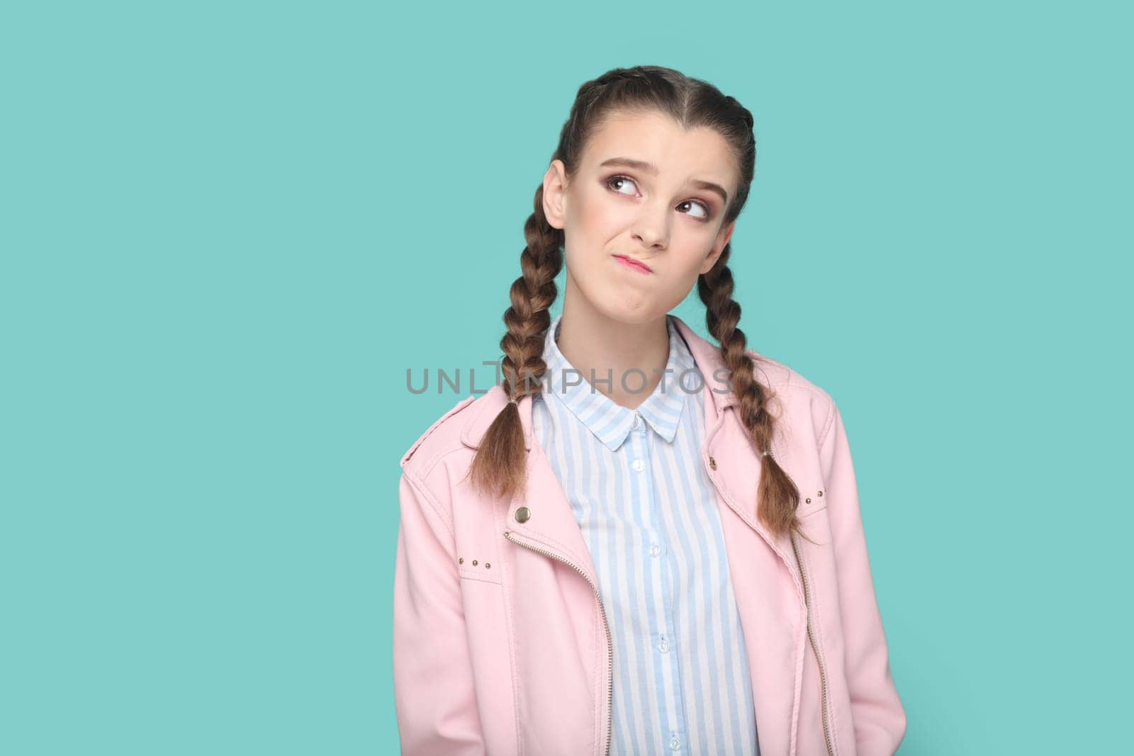 Portrait of pensive thoughtful young teenager girl with braids wearing pink jacket looking away with puzzled confused expression. Indoor studio shot isolated on green background.