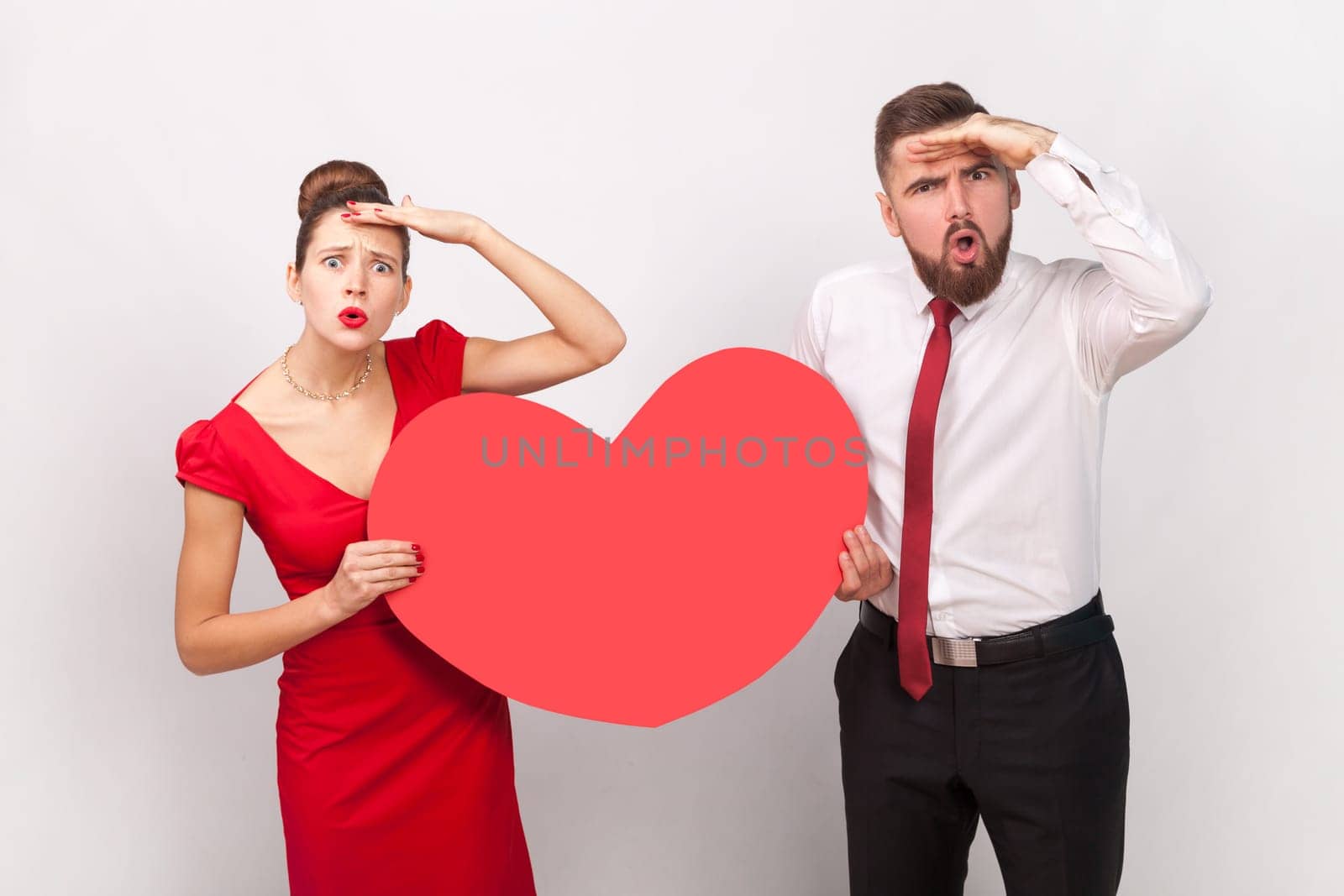 Portrait of surprised man in white shirt and woman in red dress standing together, holding big heart, looking far, waiting holiday. Indoor studio shot isolated on gray background.