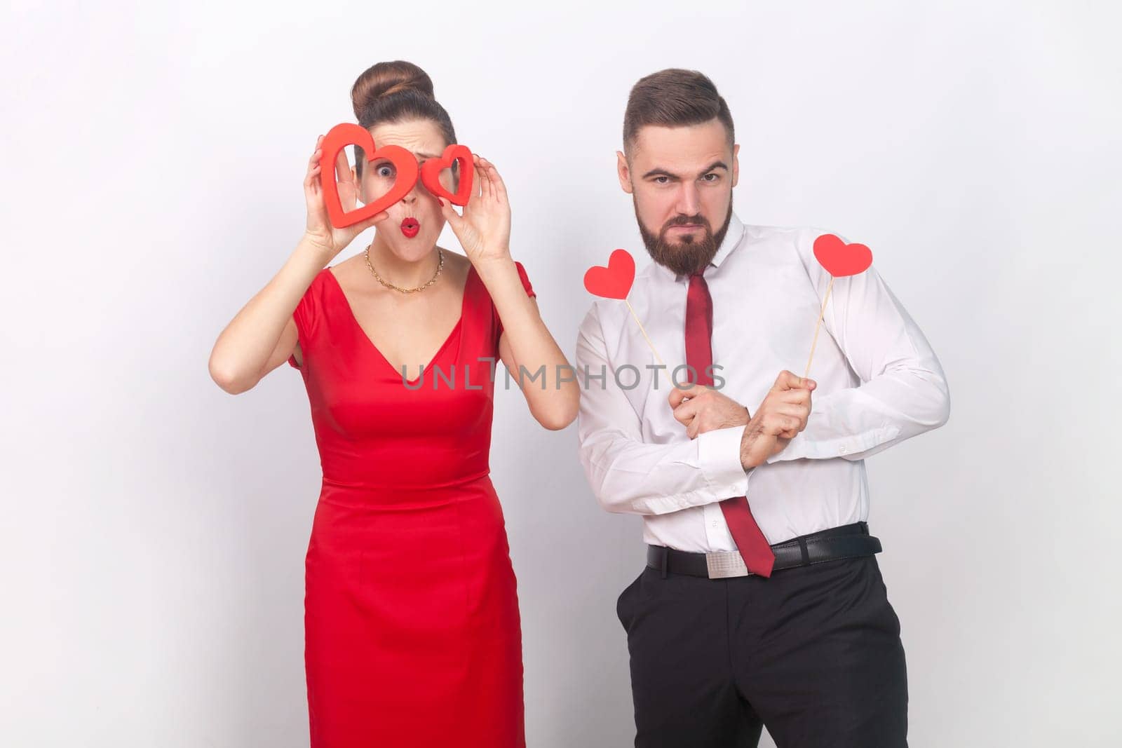 Portrait of strong confident man in white shirt and surprised woman in red dress standing together, holding heart figures, being emotive. Indoor studio shot isolated on gray background.