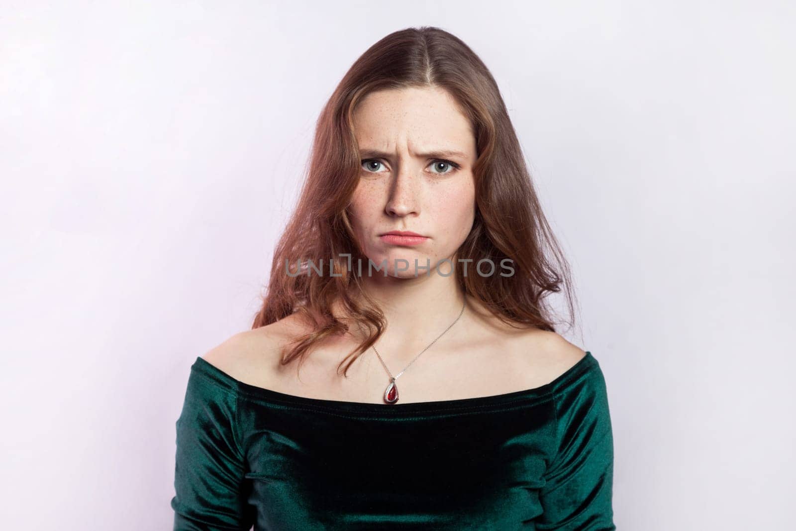 Portrait of sad worried woman wearing green dress standing looking at camera with pout lips, expressing sorrow and sadness, being offended. Indoor studio shot isolated on gray background.
