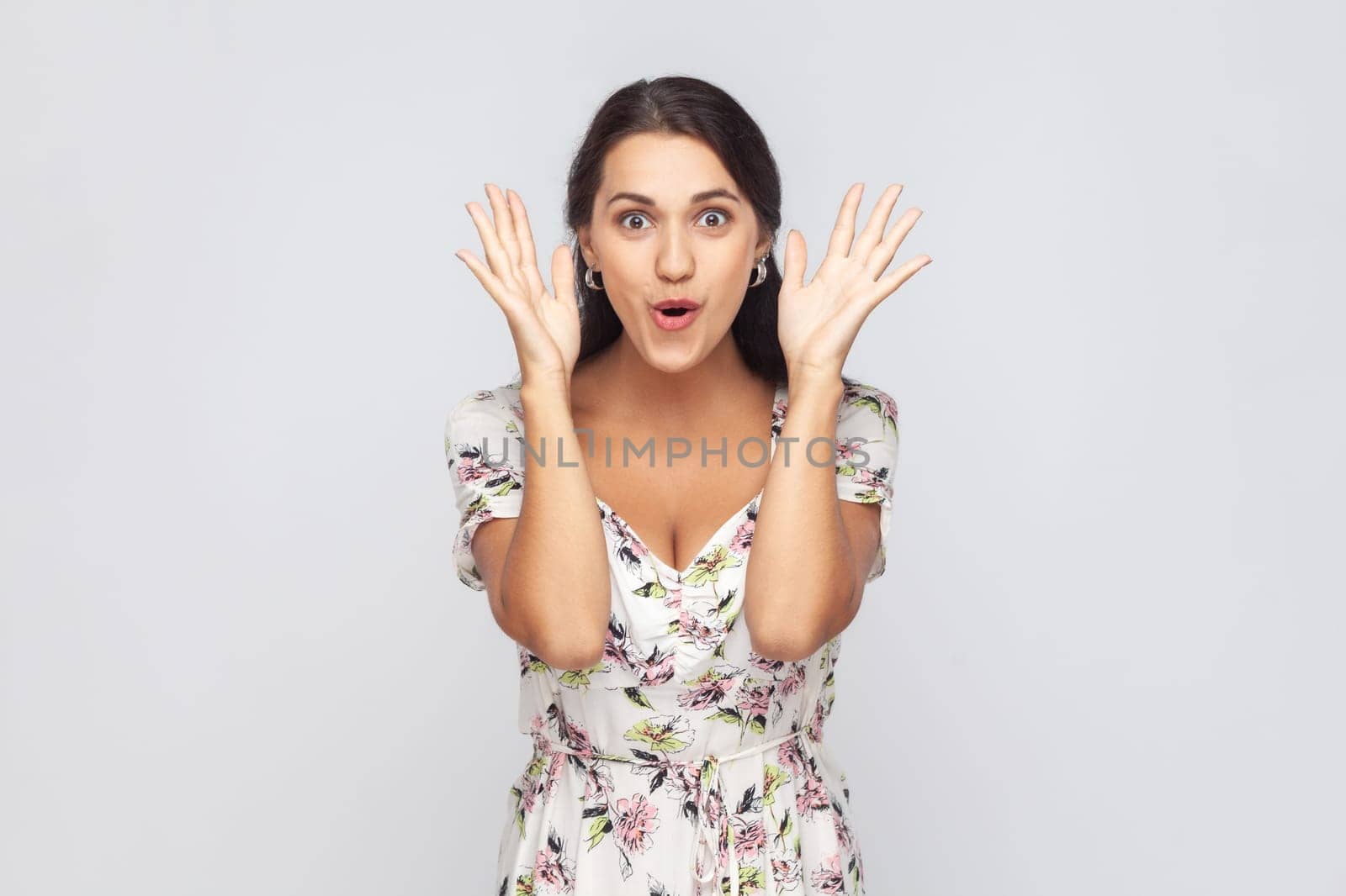 Portrait of astonished woman wearing white dress standing with mouth open in surprise, has shocked expression, hears unbelievable news, raised hands. Indoor studio shot isolated on gray background.