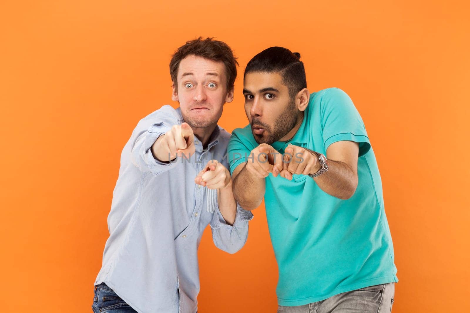 Portrait of two comic amazed men friends standing and pointing at camera, choosing you, having finny surprised expressions. Indoor studio shot isolated on orange background.