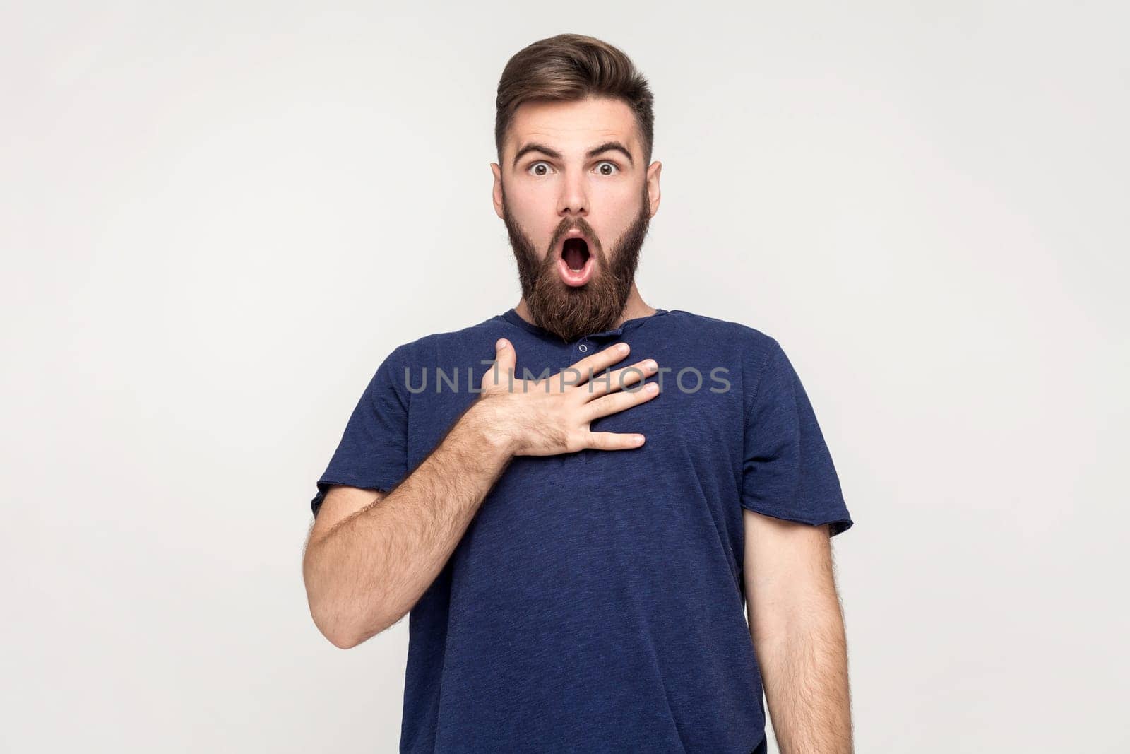 I can't believe this. Oh my god, WOW. Portrait of shocked man with beard wearing dark blue T-shirt standing and looking at camera with big eyes. Indoor shot isolated on gray background.