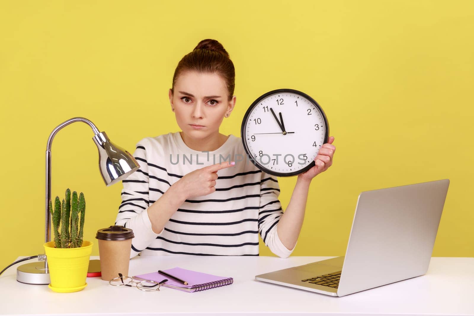 Serious bossy woman office worker pointing finger at big wall clock, time management, looking at camera with strict expression. Indoor studio studio shot isolated on yellow background.