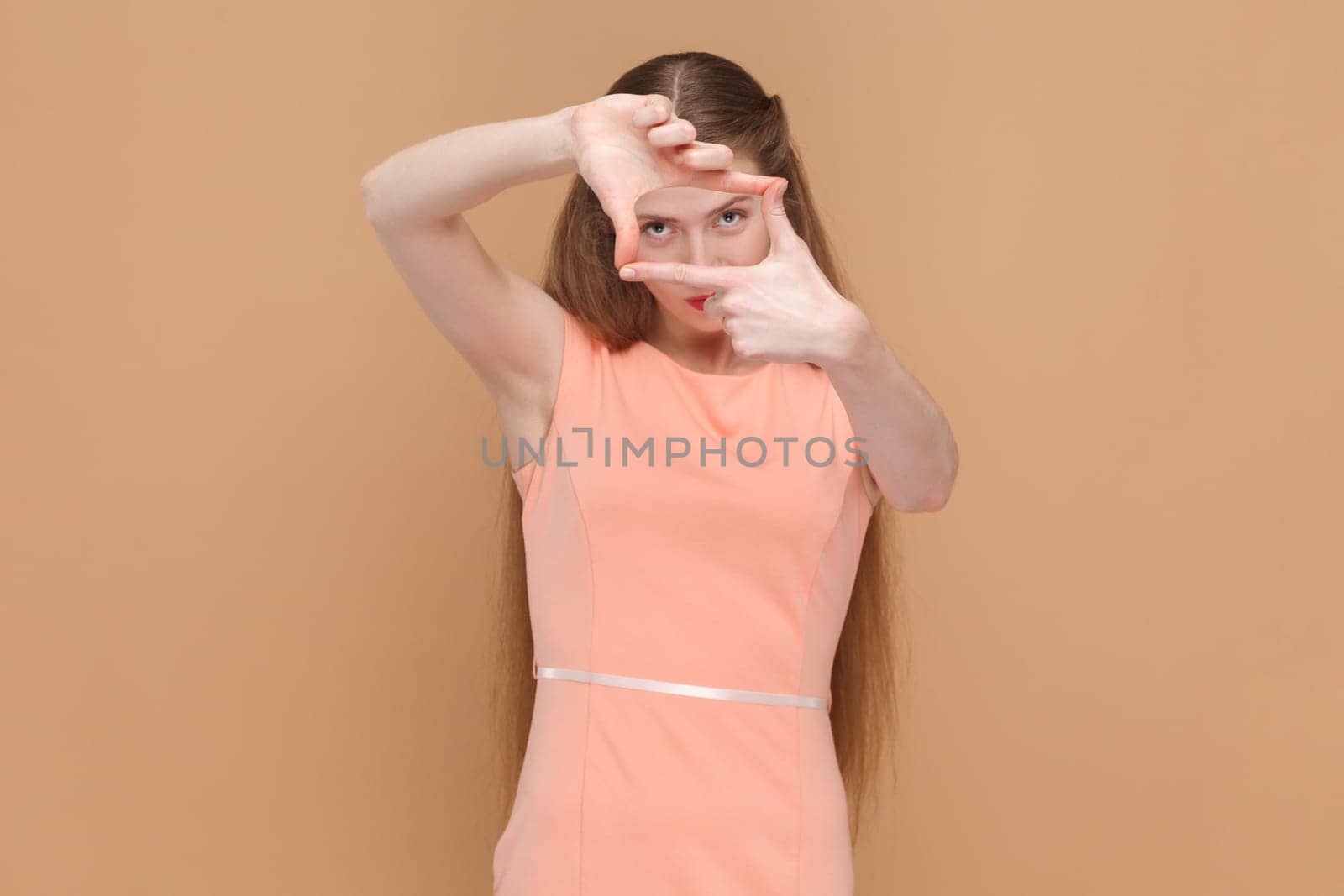 Portrait of focused attractive beautiful woman with long hair standing looking through frame made with her fingers, wearing elegant dress. Indoor studio shot isolated on brown background.