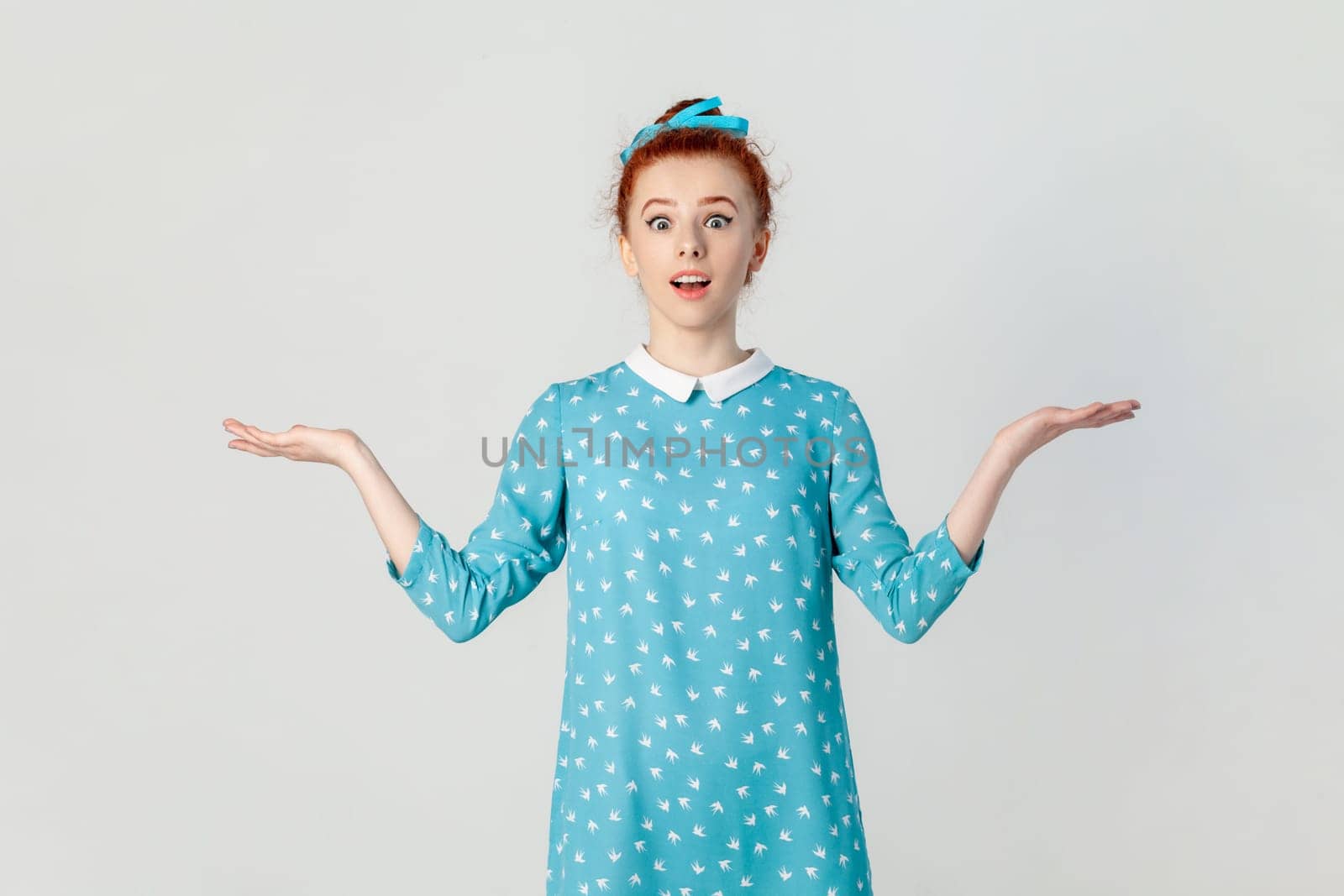 Portrait of shocked confused ginger woman standing with spread hands, looking at camera with big eyes, dosen't know how to react, wearing blue dress. Indoor studio shot isolated on gray background.